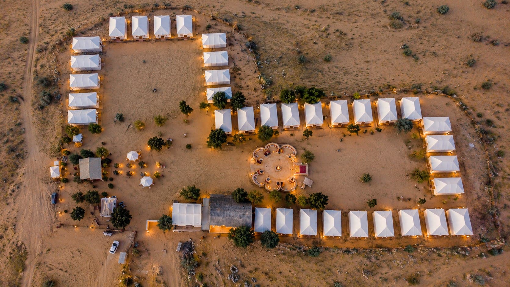 Aerial view of a desert camp at dusk: rows of white tents around an open courtyard, with a circular seating area around a fire pit and lights along the paths