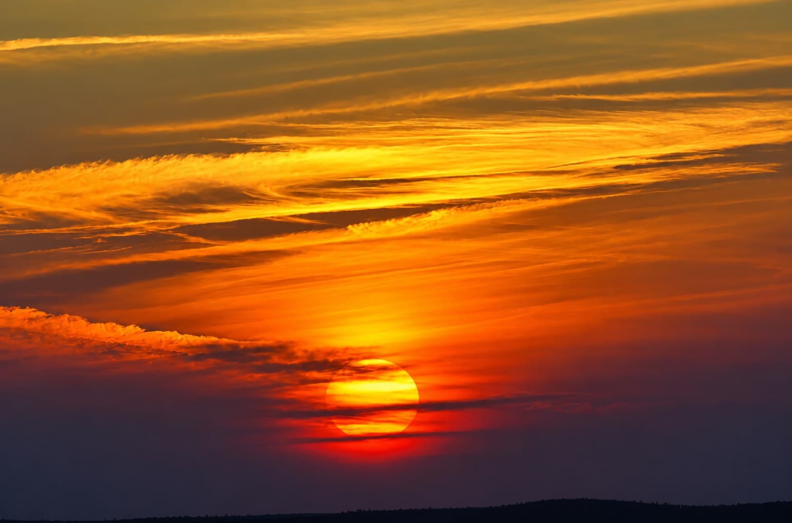 The sun setting through streaks of orange cloud above a low, dark horizon