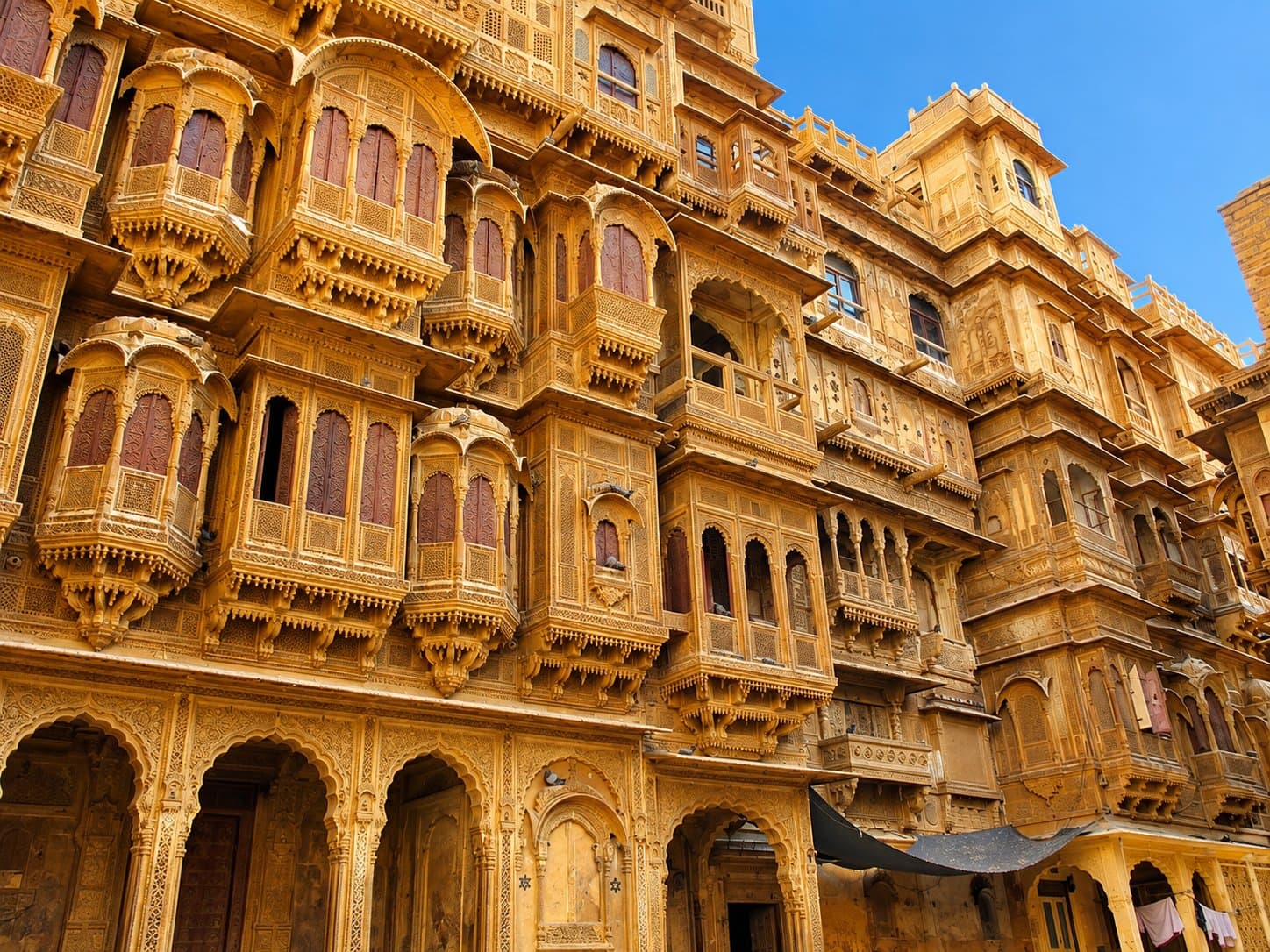 The golden sandstone facade of Patwon Ki Haveli, covered in intricately carved jharokha balconies, against a blue sky