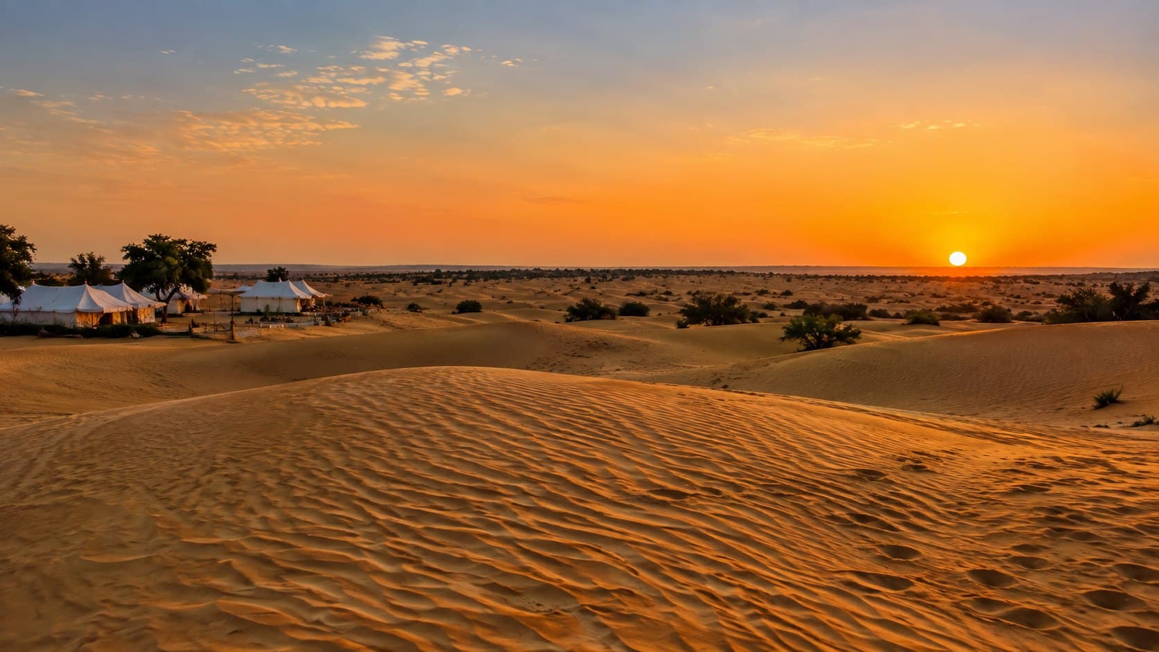 Rippled sand dunes at sunset, with the white tents of a desert camp among the trees