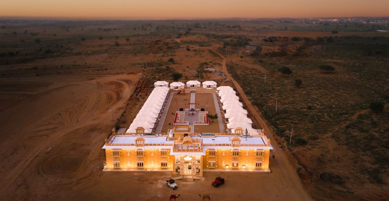 A desert resort near Jaisalmer seen from above at dusk: rows of white tents around a courtyard behind a lit, palace-style main building, with jeeps and camels at the entrance