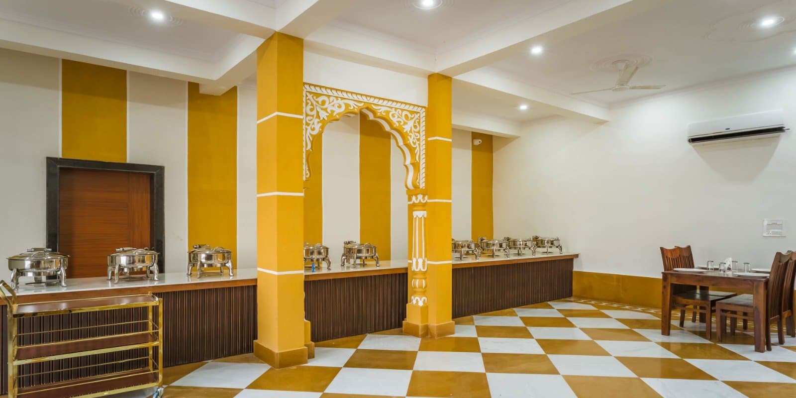 The resort’s dining hall: a long buffet counter under a carved arch, with ochre-and-white walls, a checked marble floor and wooden tables
