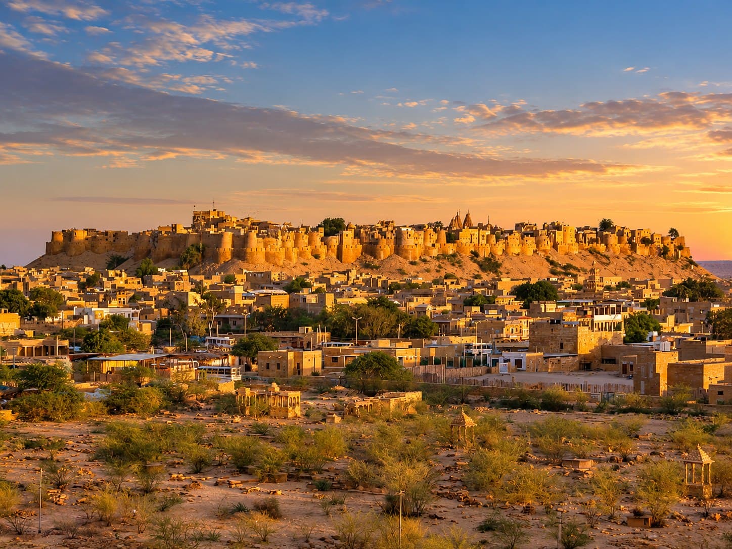 Jaisalmer Fort glowing gold on its hill above the sandstone town at sunset, with desert scrub in the foreground