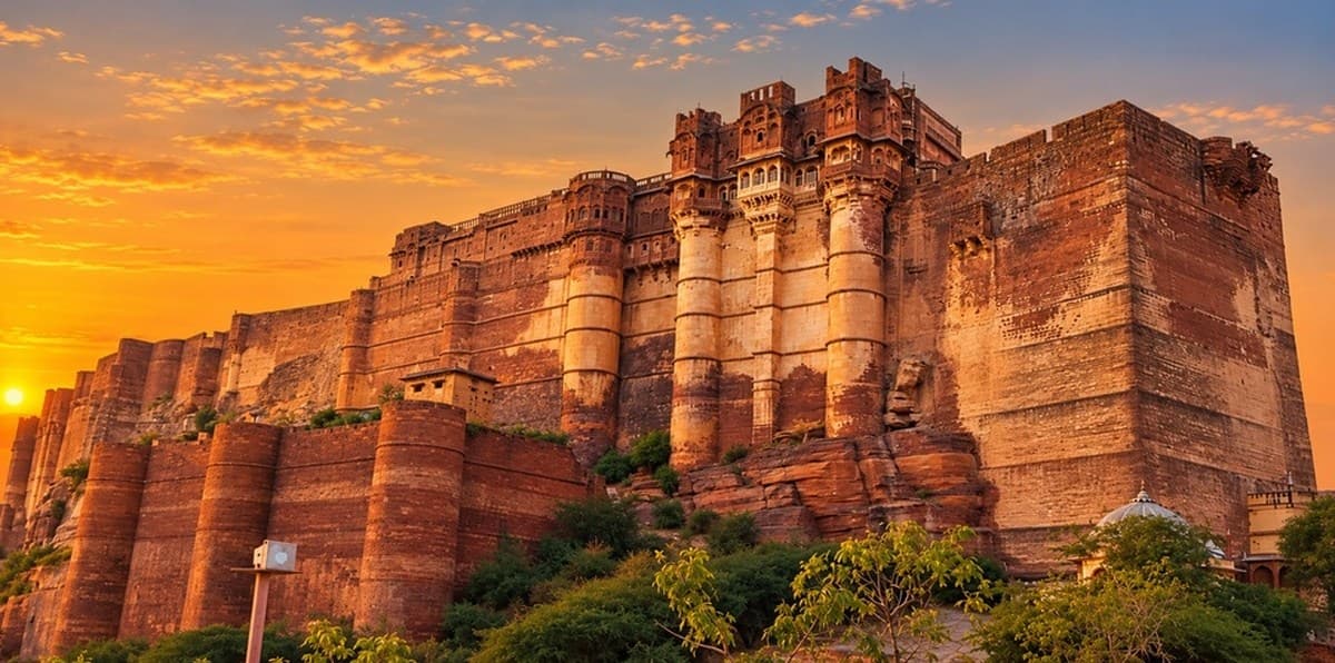 Mehrangarh Fort in Jodhpur at sunset: massive sandstone walls and round bastions rising from a rocky ridge above green trees