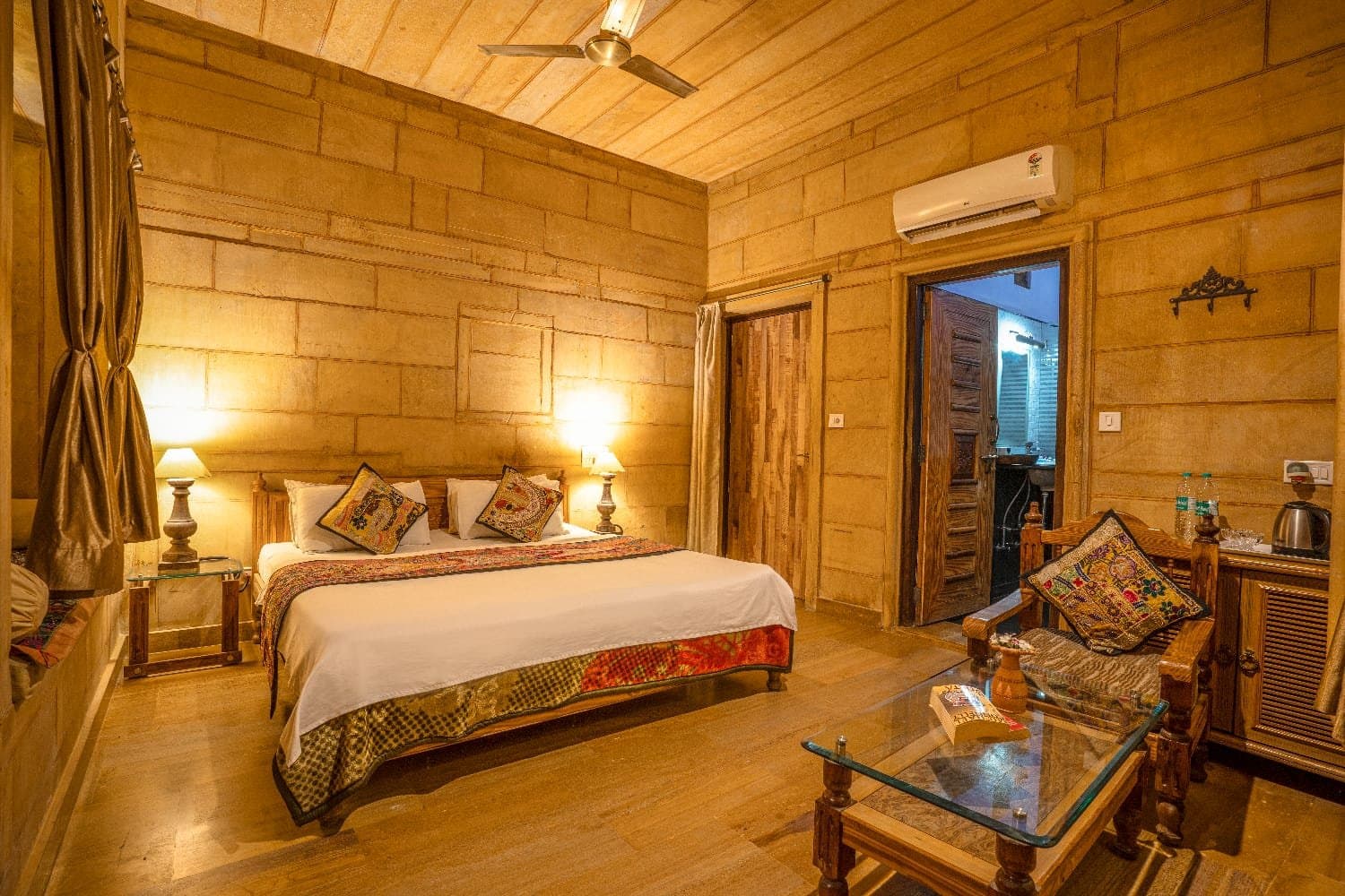 A hotel room with golden sandstone walls: a double bed with embroidered cushions, bedside lamps, a carved wooden armchair and a glass coffee table