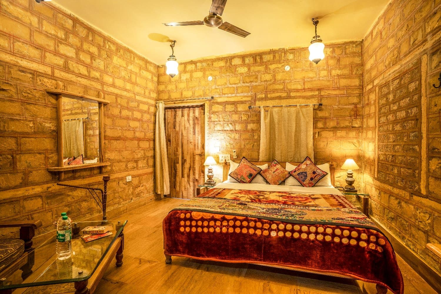 A sandstone-walled room with a double bed under a patterned red throw, hanging lanterns and a wood-framed mirror