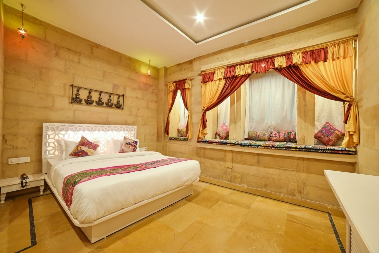 A bright room with a white carved headboard, a double bed and a long cushioned window seat under red and gold curtains